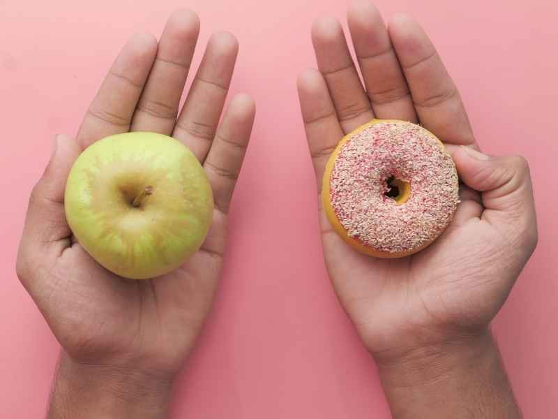 Hands holding an apple and a donut side by side, emphasizing the contrast between healthy and ultra-processed foods and the cost barriers families face.