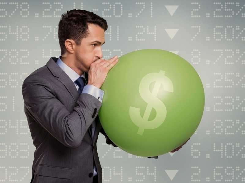 Businessman inflating a green balloon with a dollar sign, representing rising inflation and living costs.